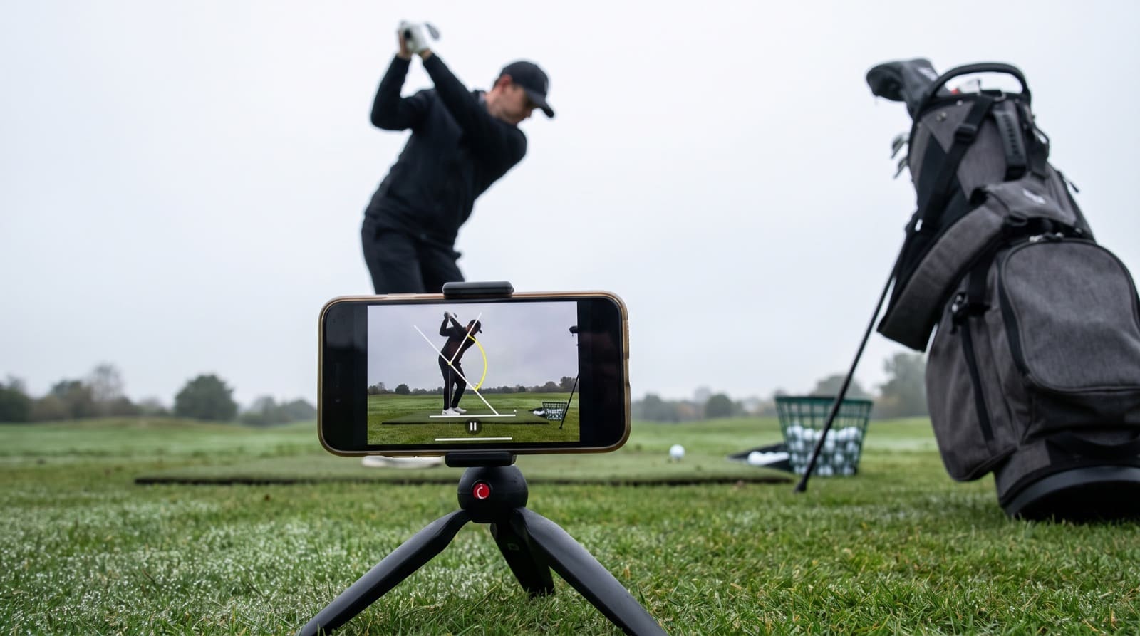 A phone on a tripod on driving range turf filming a golfer down-the-line at the top of the backswing, its screen showing the swing paused with white alignment lines and a yellow angle arc drawn over the body