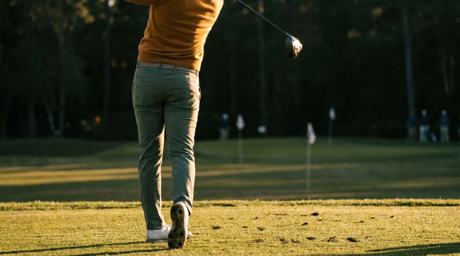 A down-the-line view of a golfer's lower body mid-swing on a sunlit fairway, hips rotated toward the target and the trail heel lifting off the turf as pressure moves into the lead foot.