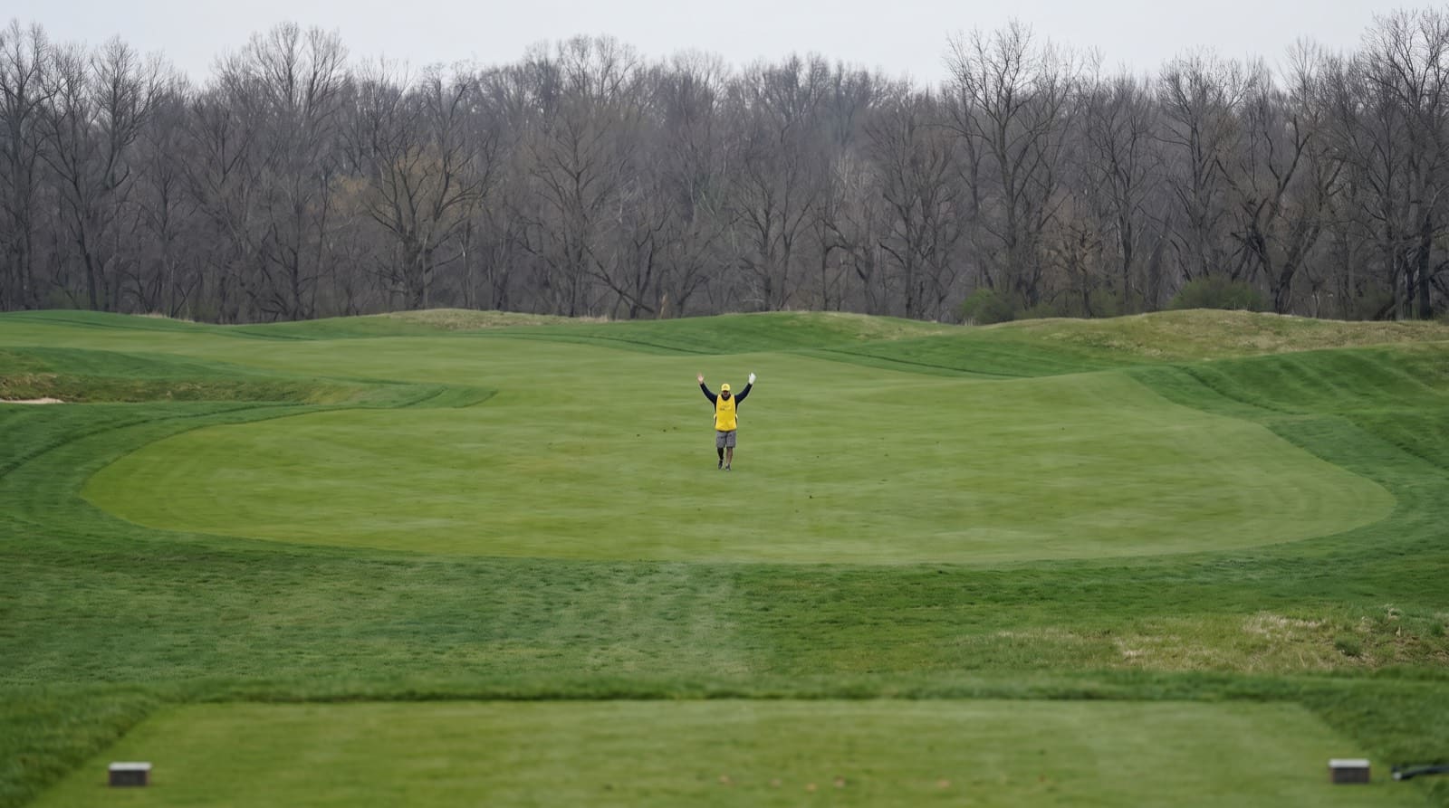 A forecaddie standing in a fairway landing zone giving the two-arms-up fairway signal back toward the tee.