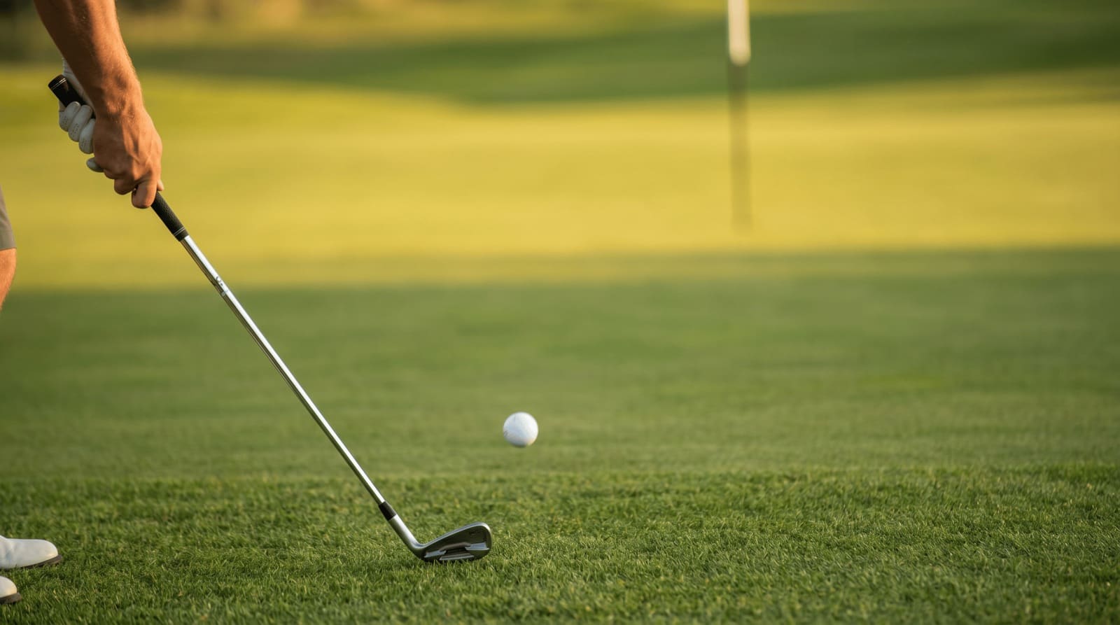 A golfer's iron brushing tight fairway turf just past a plain white golf ball, the ball launching low and starting a long roll toward a green — the minimum-air, maximum-ground bump and run.