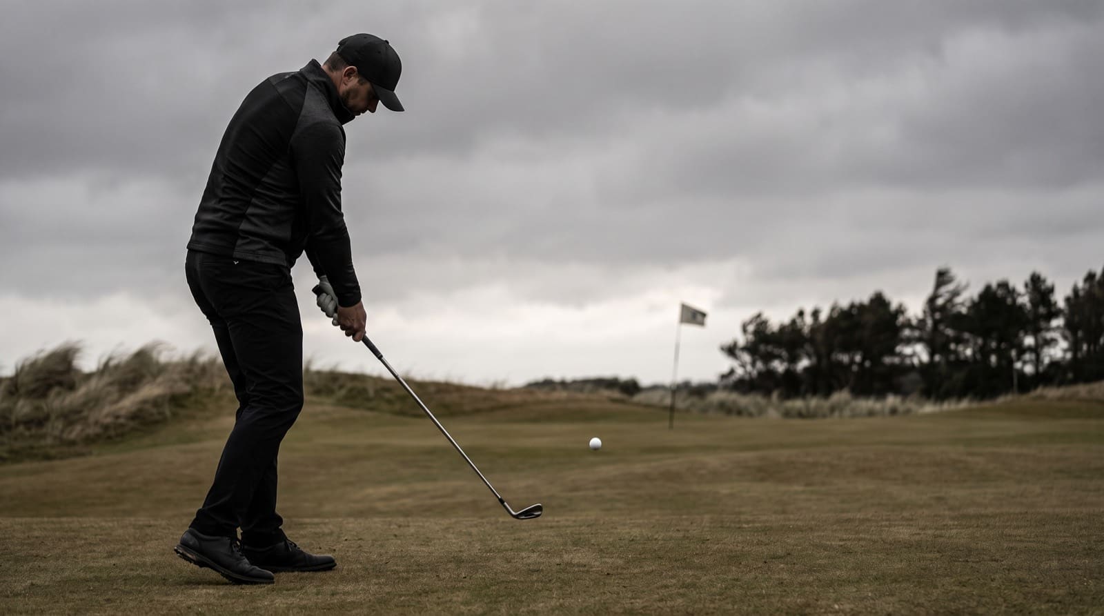 A golfer just past impact with a long iron on firm, links-style turf, the clubhead low to the ground, having struck a stinger whose ball bores away on a low, flat, penetrating line toward a distant pin, staying under the treeline beneath a heavy, windswept overcast sky.