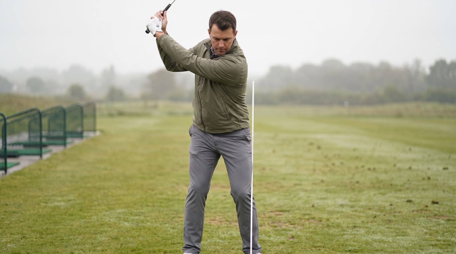 A golfer at the top of the backswing on a practice range, viewed face-on, with a vertical reference line beside the hips for checking whether the lower body has swayed off the ball.