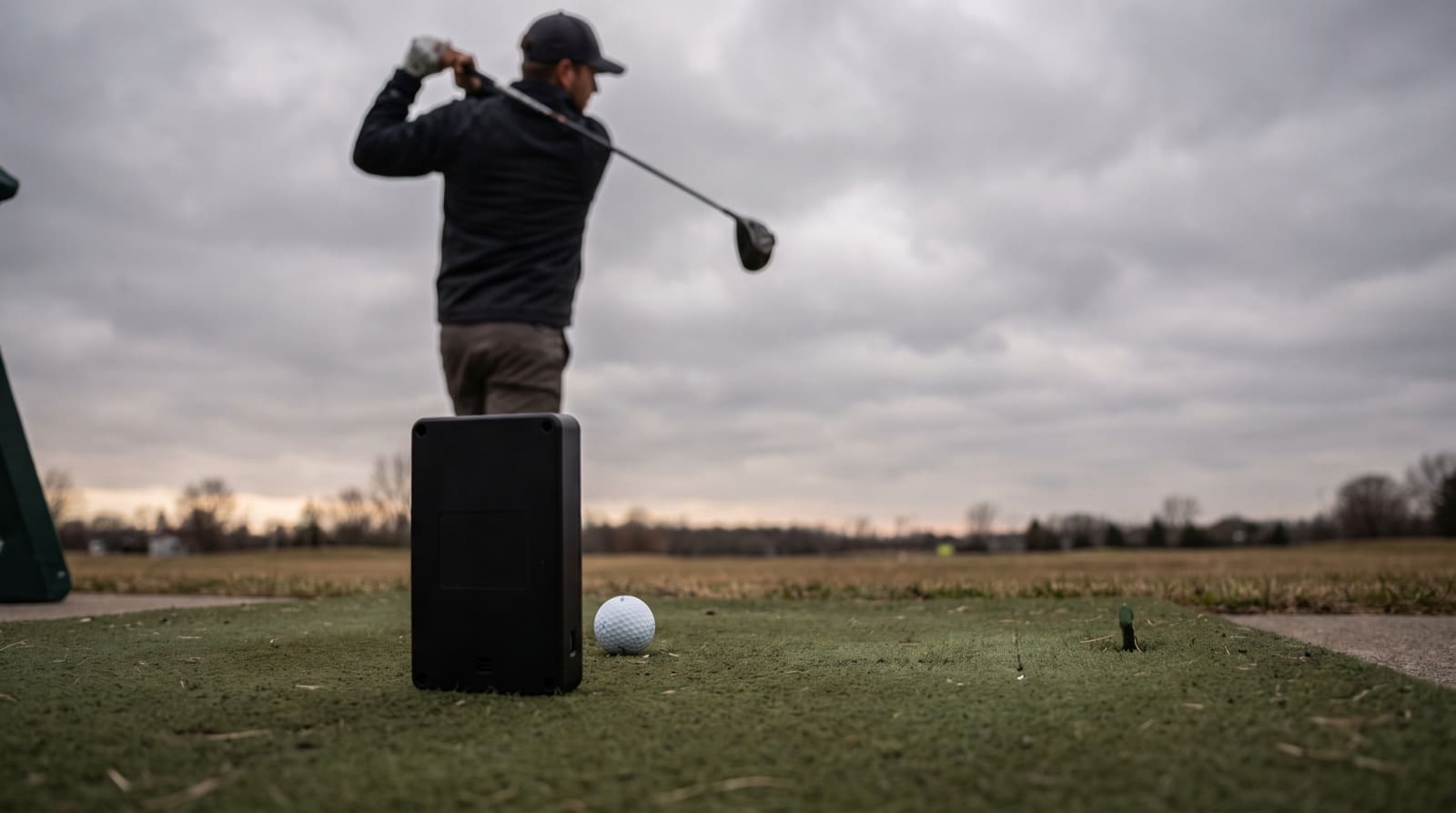 A small black radar launch monitor standing on a driving range mat directly behind a golf ball, sharp in the foreground under overcast light, with a golfer blurred in the background at the finish of a driver swing.