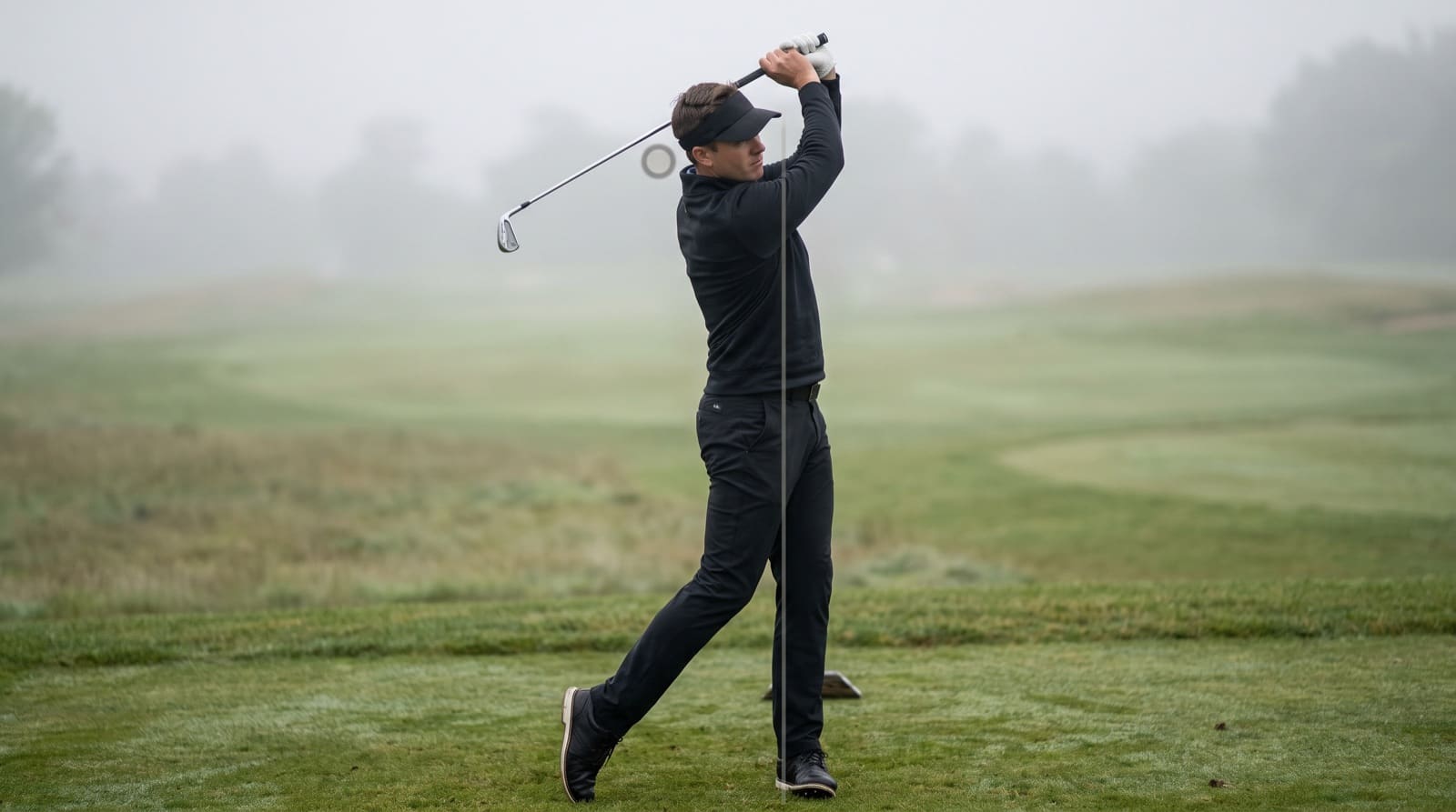A golfer at the top of the backswing on a misty course, with a vertical reference line and head-position marker used to check the pivot and weight shift.