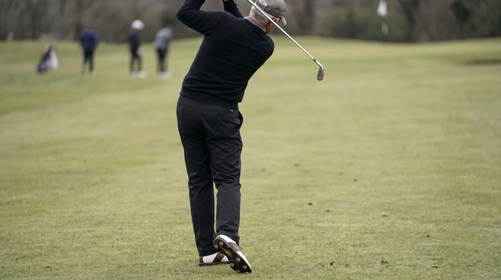 A grey-haired golfer seen from behind on an overcast fairway, swinging an iron through impact with his trail heel lifted clear of the turf and the spikes showing, hips rotated toward the target.