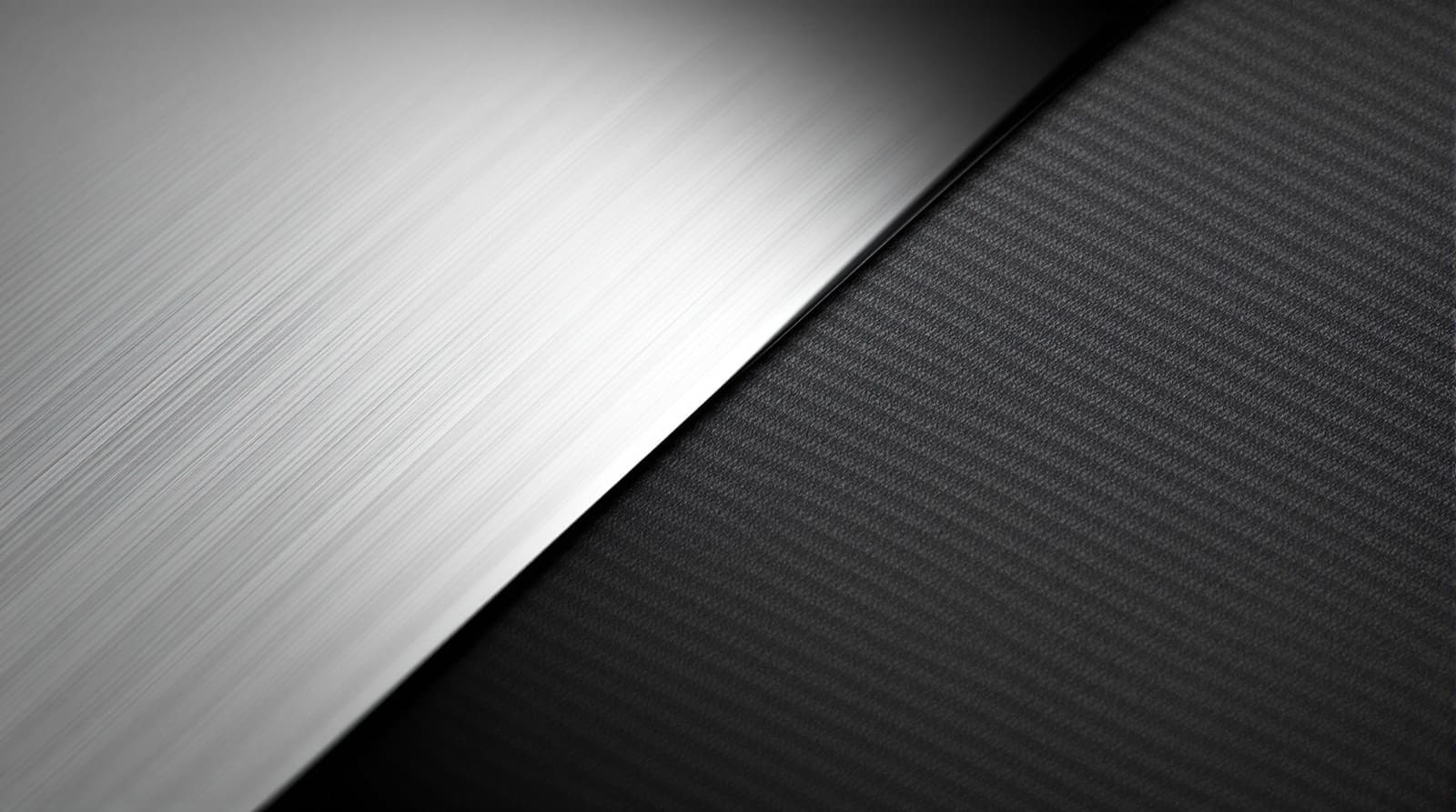 Abstract macro of two materials meeting along a clean diagonal: brushed mirror-polished steel catching a soft highlight on the left, matte black carbon fibre with a fine diagonal woven twill on the right, lit low and raking against a dark neutral ground.