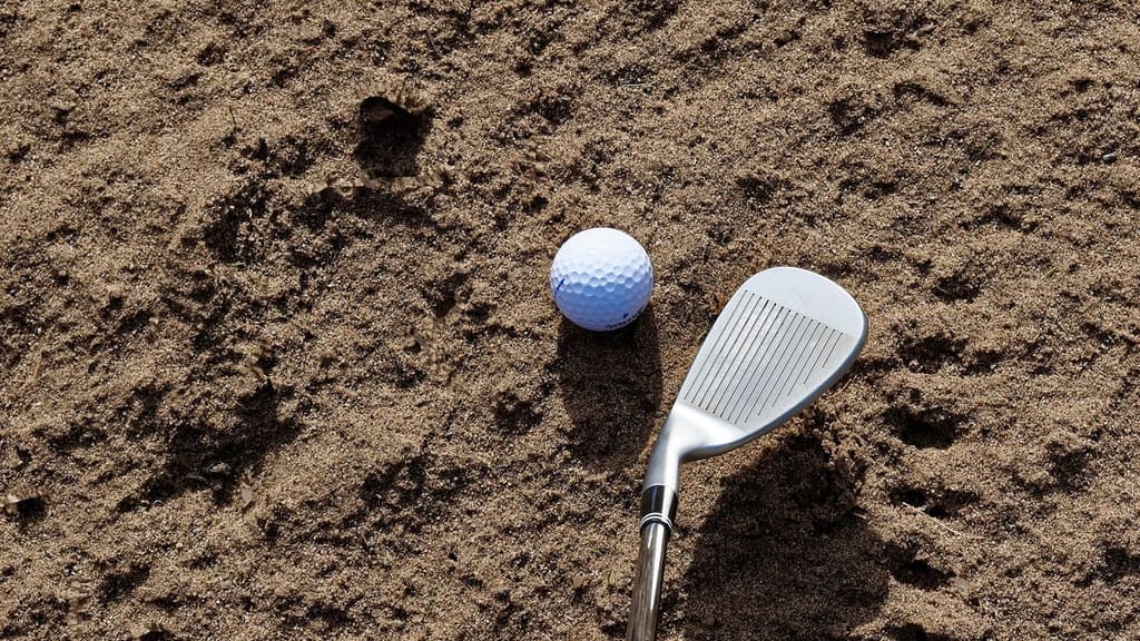 A real sand wedge resting in a bunker beside a golf ball, its tall rounded face and dense full-face grooves catching the sun, with footprints and rake marks across the coarse sand around it.